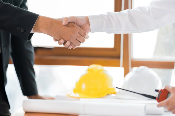 两名亚洲男性工程师或建筑师在办公室内穿着制服握手的特写镜头。工业设计概念概念。桌子上放着蓝图，头盔，对讲机。图片下载