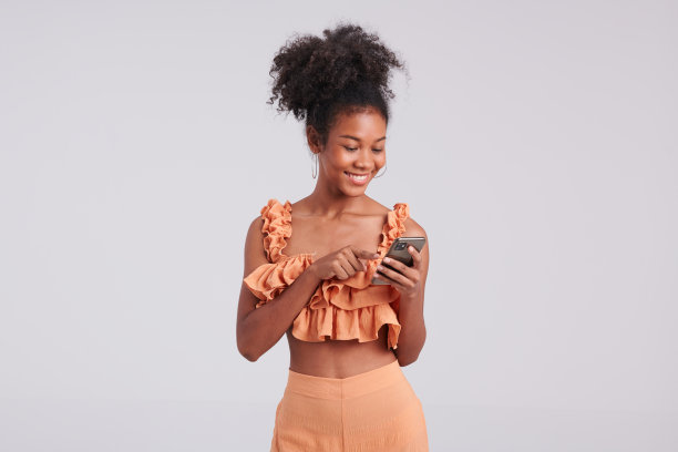 屏幕上的时尚达人:穿着橙色衬衫的黑人女孩用智能手机在工作室拍摄图片下载