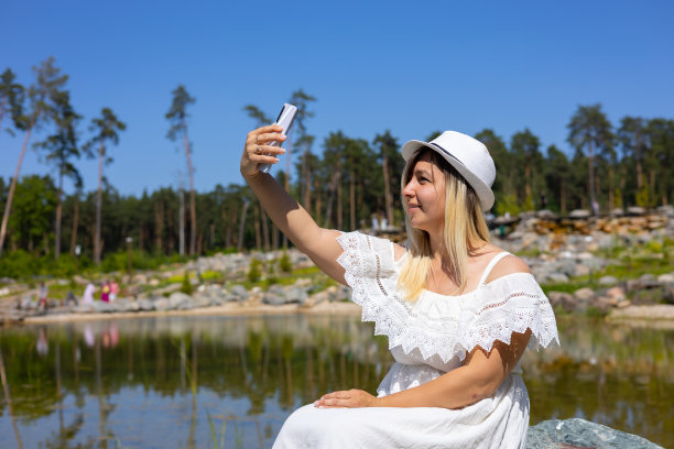 一个可爱的女人坐在美丽的湖边，用手机自拍。在一个夏日，一个穿着裙子的女人在湖边的背景下用手机拍照。图片下载