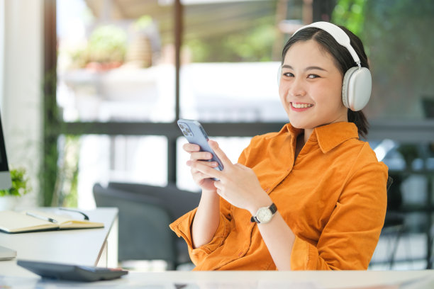 漂亮的年轻女子戴着无线耳机在明亮的现代办公室里使用手机。图片下载