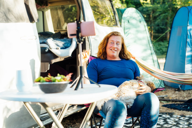 中年游牧妇女在森林营地的露营车旁与宠物狗坐在一起打视频电话。旅行博主在自驾游假期工作。与关注者在线交流图片下载