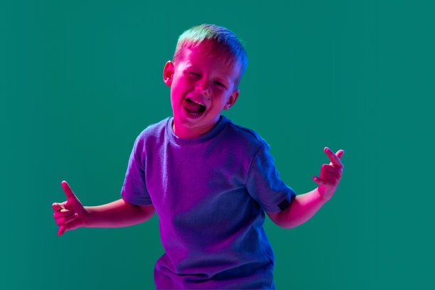 一个顽皮的小男孩的肖像，孩子穿着休闲的衣服玩耍，在霓虹灯下的青色工作室背景下露出有趣的脸图片下载