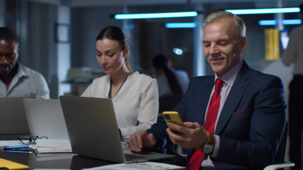 成熟的商人在笔记本电脑上工作，微笑着在智能手机上阅读文本图片下载