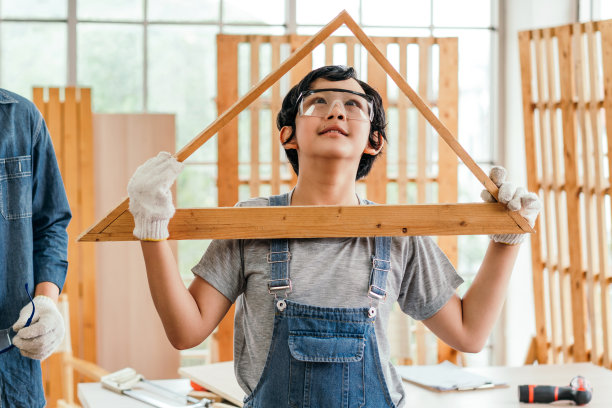 图为一个欢快的亚洲家庭，木匠儿子戴着护目镜和手套，手持自制的手工三角形木结构，站在家里的木制工作室里图片下载