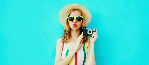 夏天的肖像快乐时尚的年轻女性摄影师与电影相机吹她的嘴唇与红色唇膏戴草帽在蓝色的背景，空白拷贝空间的广告文字图片下载