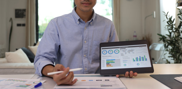 年轻的商人坐在办公桌旁，看着相机，指着一个平板电脑，上面显示着图形形式的财务信息(柱状图、堆叠条形图和饼状图)。图片下载