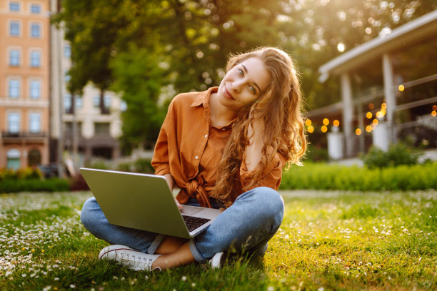 美丽的自由职业者坐在绿色的草地上，手里拿着一台笔记本电脑。图片下载