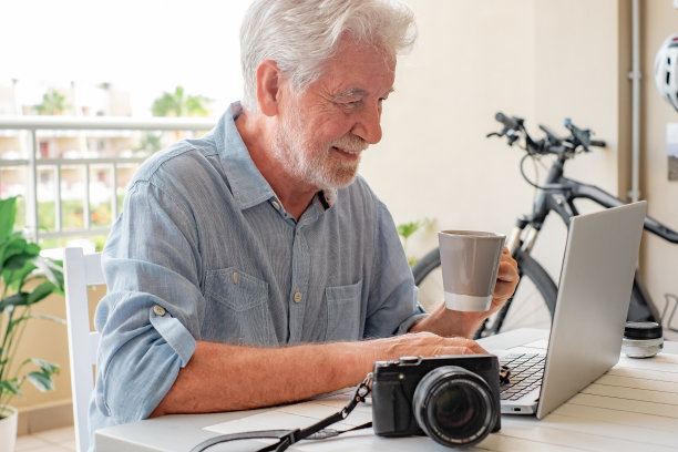 快乐的白发老人坐在户外露台上拿着咖啡杯，在笔记本电脑键盘上打字，老人在家用网络技术做生意图片下载