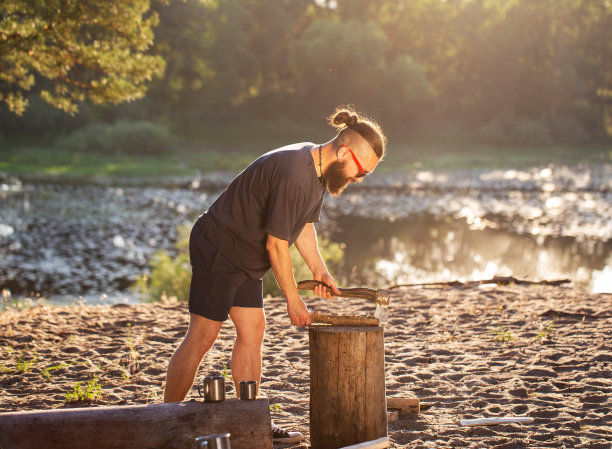 一个留着胡子，手里拿着斧头的野蛮白人男子正在露营地砍柴生火。夏天在河边的户外娱乐。Bushcrafting。日落图片下载