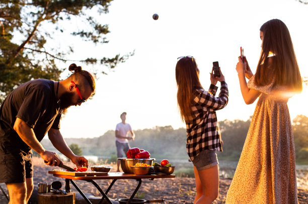 一个留着胡子、戴着黑色眼镜的野蛮人，在夕阳的背景下，正在大自然中准备一份沙拉。女生用手机拍照，男生打排球。夏季户外娱乐。文本复制空间图片下载
