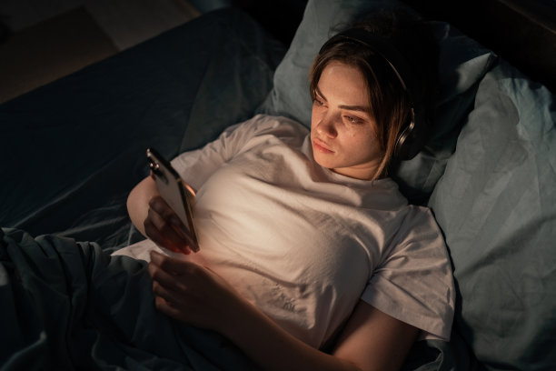 年轻疲惫的女子在睡前戴着耳机听音乐和朋友聊天，饱受失眠之苦。晚上聊天和冲浪电话。社交网络和手机成瘾。图片下载