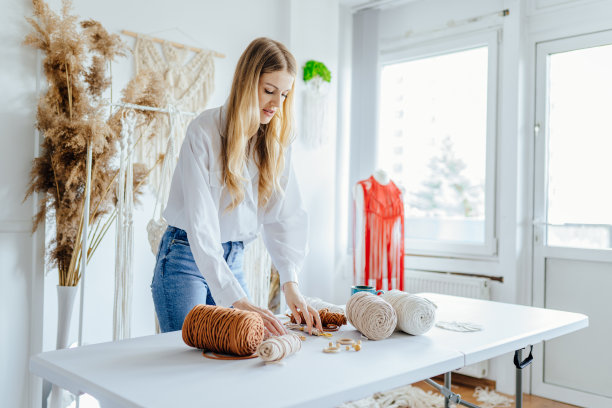 金发美女在她舒适的小工作室里从事创作。手工编织带在工作过程中。女性的爱好。图片下载