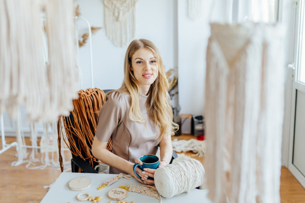 友好的女工匠在makrame工作室喝茶的肖像。在编织大师班或手工艺品店工作后喝咖啡休息。女艺术家坐在她手工制作的makrame周围的工作区。图片下载