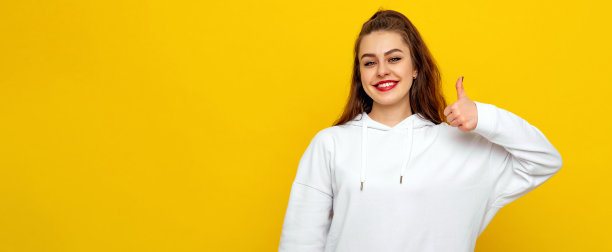 快乐漂亮的年轻黑发女子穿着白色休闲风格的运动衫，笑着竖起大拇指。室内工作室拍摄的黄色背景与文本拷贝空间图片下载