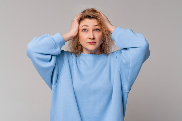 金发压力中年妇女的肖像手放在头上，穿着时髦的蓝色运动衫，站在灯光工作室背景图片下载