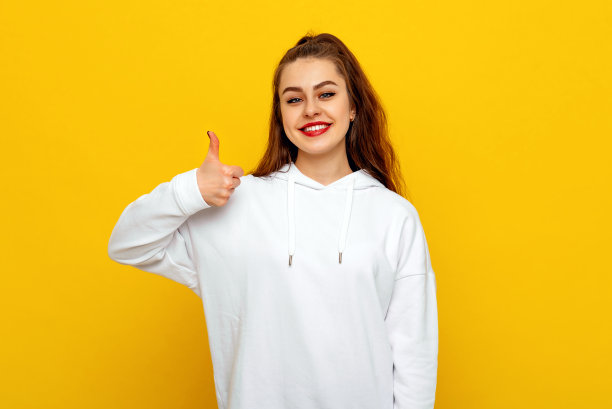 自信漂亮的年轻黑发女人在白色休闲风格运动衫，微笑和显示大拇指。室内工作室拍摄黄色背景图片下载