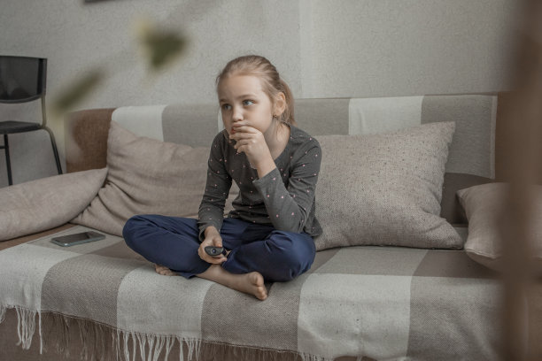 一个可爱的小女孩正坐在家里的沙发上，用遥控器切换电视频道。为儿童和青少年观看内容丰富、有趣的儿童节目或视频。图片下载