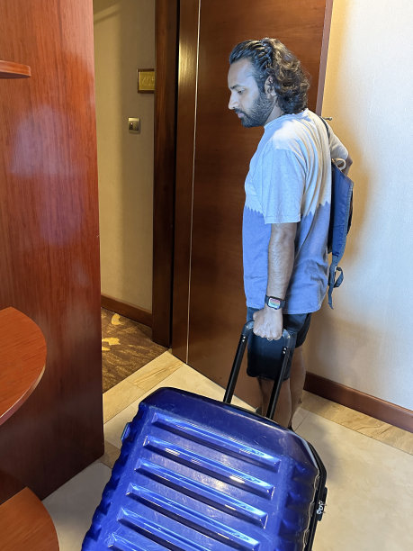 酒店客人背着帆布背包，拖着滑轮行李箱，离开酒店的特写形象，打开的木门，走廊棕色和金色，抽象有趣的图案地毯，重点放在前景图片下载