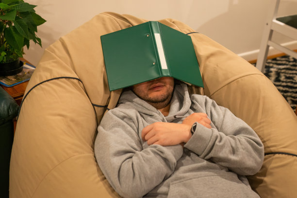 一个年轻人躺在豆袋上，脸上蒙着绿色的文件夹。从学习概念中休息一下。图片下载