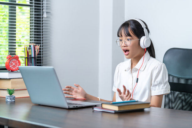 快乐微笑的女学生戴着耳机在家庭办公室用笔记本电脑在线讨论会议。学习远程教育业务的女大学生图片下载