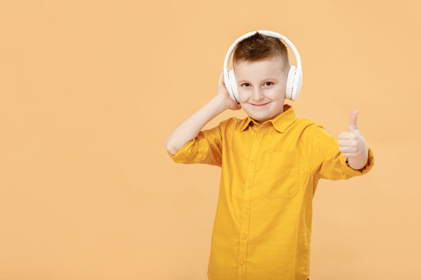 滑稽聪明的男孩肖像，戴着耳机，穿着黄色衬衫。黄色工作室背景。教育。面带微笑，对着镜头竖起大拇指。高质量的照片图片下载