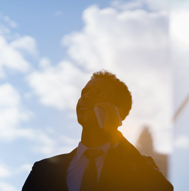 身着蓝色西装、打着领带的黑人商人的剪影，在日出或日落的光线下使用手机、打电话、在城市建筑外工作。有人在看复印空间图片下载