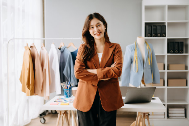 亚洲年轻女子坐在时装设计师办公室的桌子上，白色桌子上放着平板电脑、笔记本电脑和智能手机图片下载
