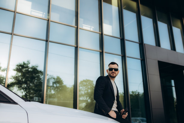 在现代办公中心附近的街道上，一个英俊、微笑、快乐、留着胡子的西装男子看了看手机，走向他的新白色汽车。图片下载