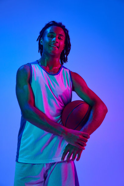 在霓虹灯的渐变蓝色背景下，一个穿着制服的篮球运动员带着球摆姿势图片下载