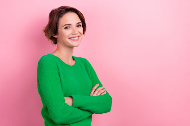 照片中，年轻迷人的商务女士穿着绿色衬衫，双手交叉，在粉红色的背景上表现出自信图片下载