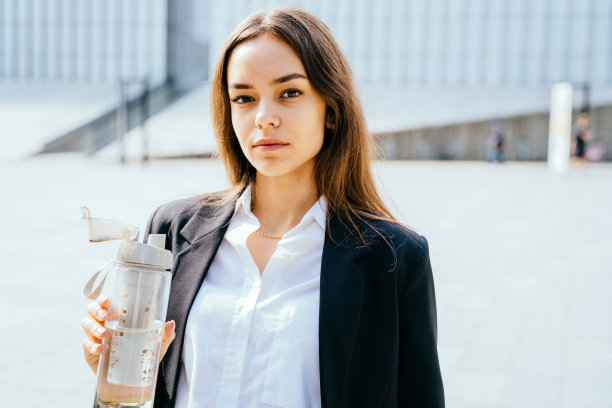 年轻女学生拿着瓶装水站在金融区户外，背景是现代建筑。图片下载