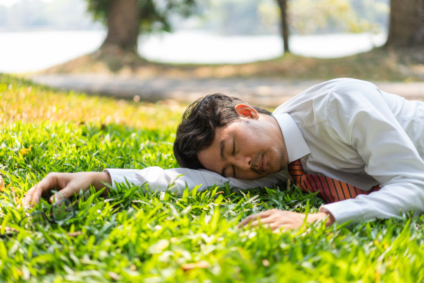 午餐时间，在附近的一个公园里，疲惫不堪的男职员戴着摘下的眼镜趴在地上，用一只手撑着头在草坪上打盹。图片下载