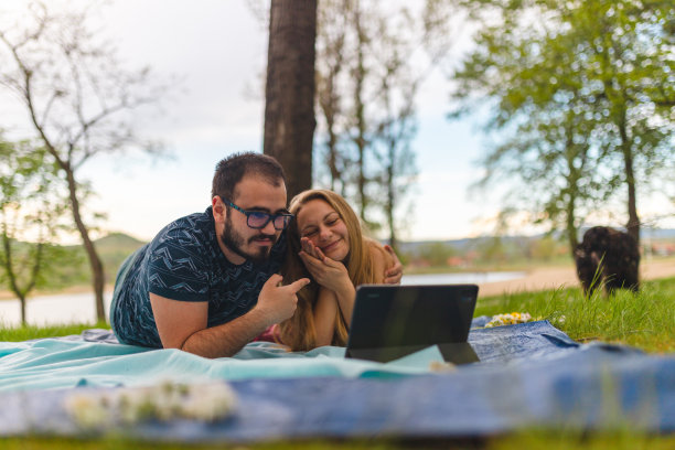 一对年轻夫妇躺在野外的野餐毯子上，同时用平板电脑进行视频通话图片下载