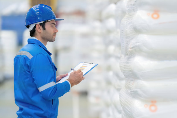 美国男工程师在大仓库的半边照片用笔核对产品订单，用清单记事，在生产塑料和钢铁材料的大型工业厂房戴着头盔，穿着制服。图片下载