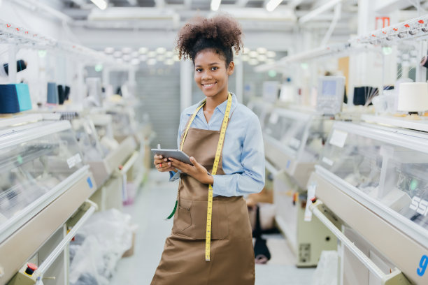 美国女设计师手持平板电脑和卷尺自信地微笑着站在纺织行业穿着制服和缝纫机周围工作图片下载