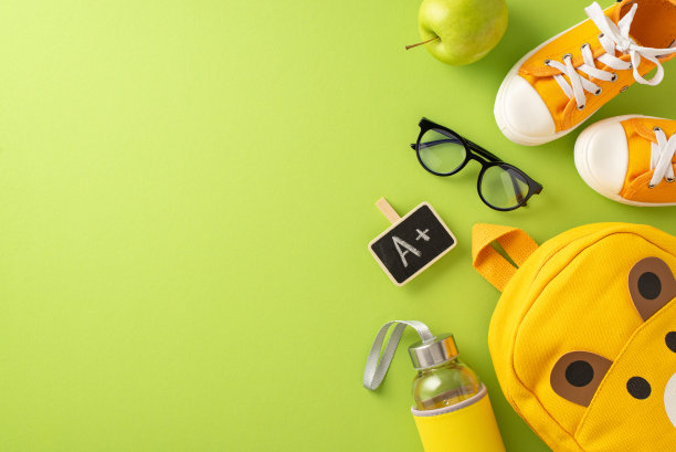 准备开始上小学。可爱的熊主题背包，A+荣誉，眼镜，运动鞋，苹果和水瓶在柔和的绿色背景，文字或广告的空间俯视图图片下载