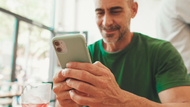 快乐的成熟男人穿着休闲的衣服，坐在咖啡馆用智能手机，看照片和视频图片下载