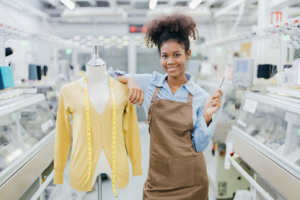 美国女设计师潇洒地站着，穿着制服，微笑着，拿着一支笔站在一个卷尺模特旁边。在织造工业中到处都是缝纫机。图片下载