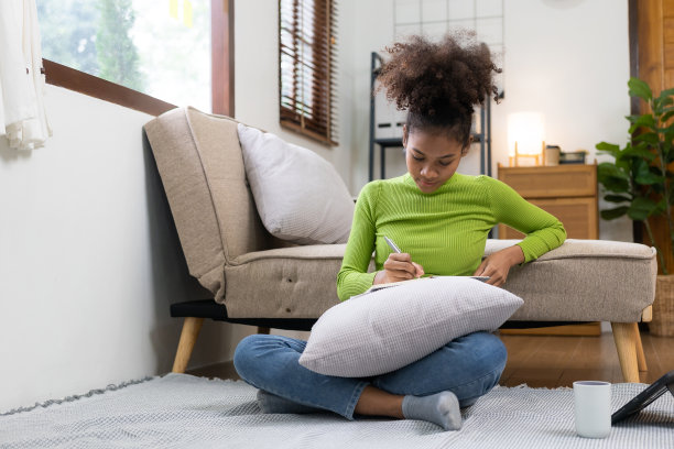 年轻的非洲妇女微笑着坐在客厅软沙发旁边的地板上使用平板电脑。在社交网络上享受网上购物。图片下载