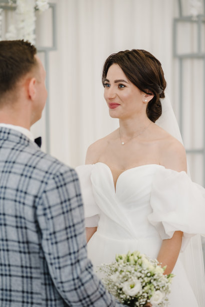 誓言和约定。新婚夫妇手持花束站在装饰着白花的拱门下举行婚礼。新娘和新郎结婚。女人和男人同时举行仪式。婚姻。婚纱照片。图片下载