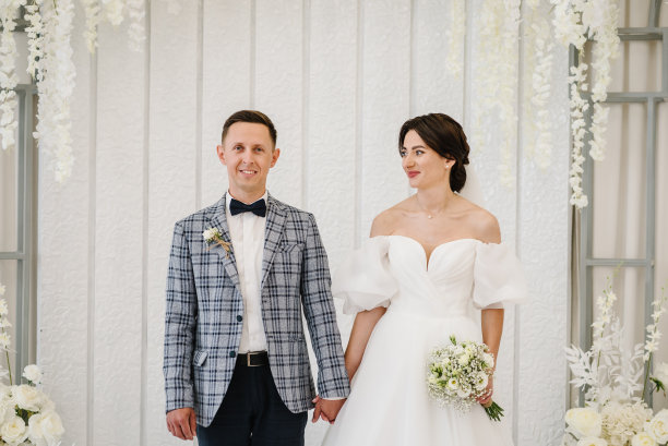 新婚夫妇手持花束站在装饰着白花的拱门下举行婚礼。新娘和新郎结婚。有魅力的女人穿着礼服和男人在仪式上。婚姻。婚纱照片。图片下载