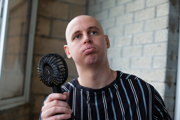 一个汗流浃背的秃顶男子正在用便携式小风扇给自己吹风降温图片下载