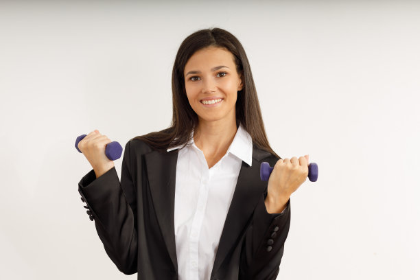 深色头发，穿着时髦的商务套装和白衬衫的女人拿着小的女性哑铃。在孤立的白色背景上，快乐微笑的年轻女子带着运动器材的肖像。图片下载