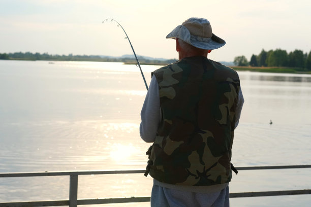 老人钓鱼。夕阳西下，一位白发苍苍的老渔夫从码头上抛下一缕丝线。积极的老年男性在湖中垂钓，旋转卷轴杆。退休渔民享受他的周末图片下载