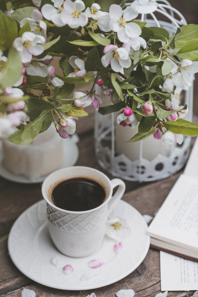 一个漂亮的明信片。一个白色咖啡杯，一个茶托，一个小雕像，蜡烛，一本书和一个插着一束盛开的苹果树的花瓶。美丽的生活。春天的时间。“早上好”的概念图片下载