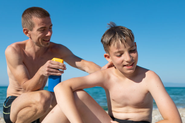 阳光明媚的日子里，父亲在沙滩上给光着膀子的儿子背上涂防晒霜。家庭，生活方式和护理。图片下载