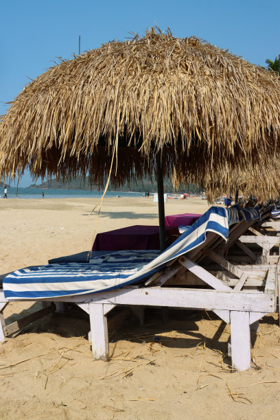 印度果阿，帕洛勒姆海滩，一排木制的蓝色软垫躺椅，在沙滩上晒日光浴，茅草遮阳伞，蓝色和白色条纹的毛巾，边桌图片下载
