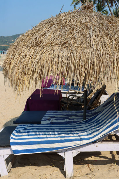 印度果阿，帕洛莱姆海滩，沙滩度假的特写照片，沙滩上有一排成对的蓝色软垫太阳躺床，茅草遮阳伞，蓝色和白色条纹的毛巾，边桌图片下载
