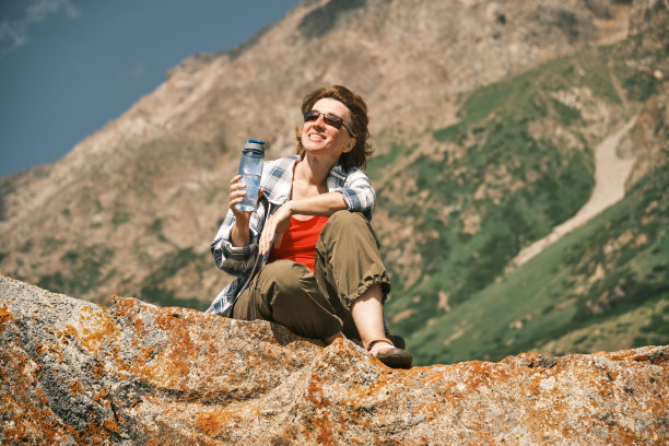 快乐的46岁女游客坐在高山上的岩石上喝水图片下载