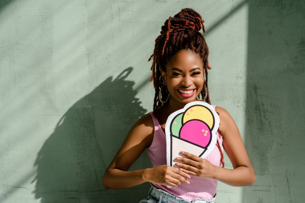 在一个阳光明媚的夏日，一个快乐微笑的年轻黑人妇女的户外肖像，她梳着脏辫，戴着粉红色的上衣，戴着圆形耳环，还有一张印有冰淇淋的照片。副本的空间。广告图片下载